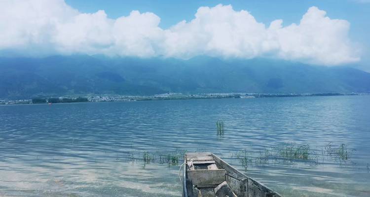 Lac bleu calme avec un bateau en bois au premier plan et des chaînes de montagnes voilées par les nuages en arrière-plan.