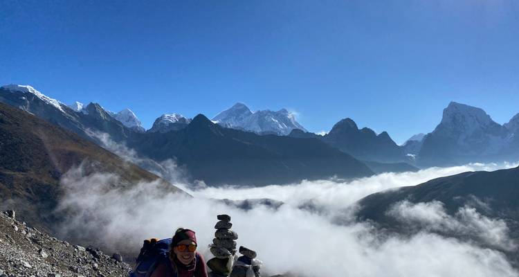 Excursionista posando junto a un pequeño mojón de piedra con vista a las nubes que flotan en un valle con picos dentados del Himalaya más allá.