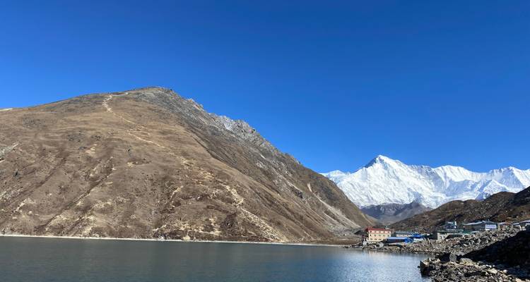 Tranquilo pueblo himalayo junto al lago enclavado entre laderas áridas y pardas y picos nevados distantes reflejados en aguas calmadas.