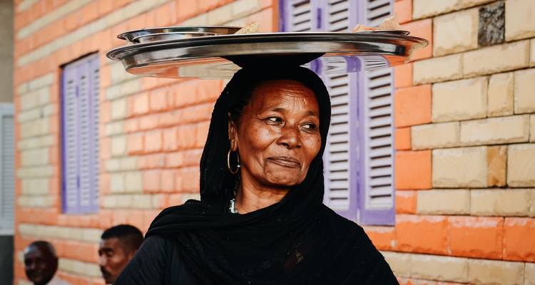 Een vrouw die een dienblad op haar hoofd draagt, staand tegen een kleurrijke muur.