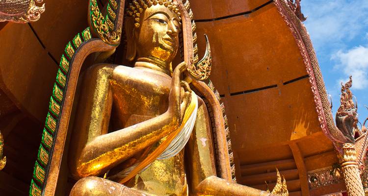 Una reluciente estatua de Buda dorado sentado dentro de un pabellón al aire libre ornamentado contra un cielo brillante.