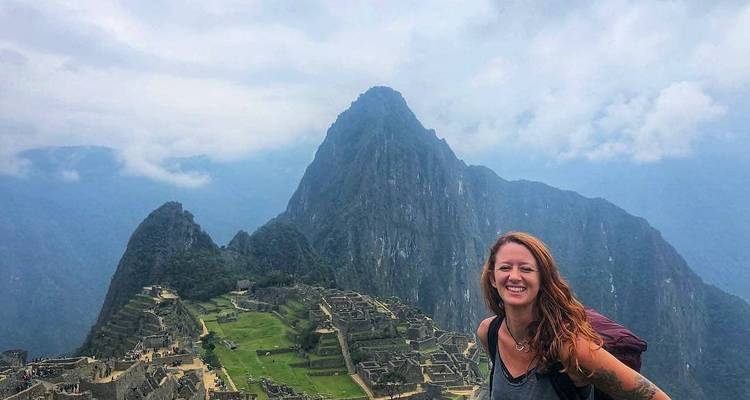 A woman smiling with Machu Picchu in the background under a cloudy sky.