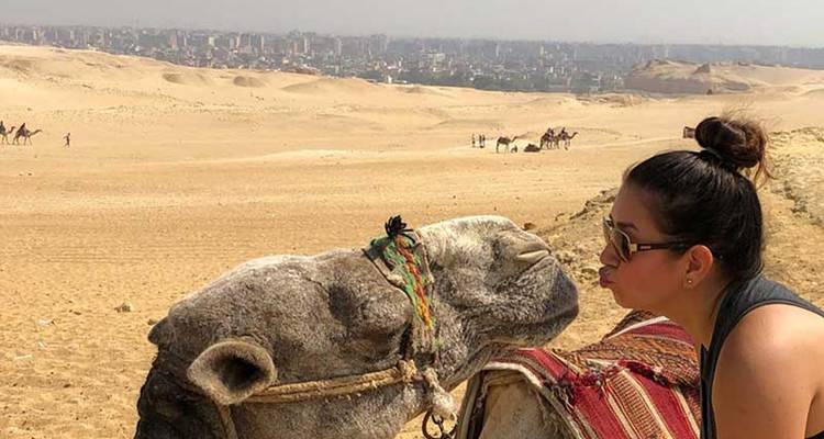 Femme posant avec un chameau dans le désert avec un paysage urbain en arrière-plan.