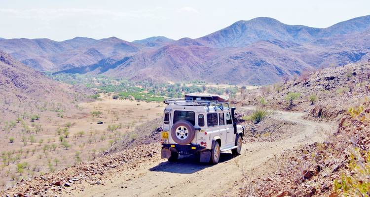 A white SUV driving along a dirt road with mountainous terrain in the distance.