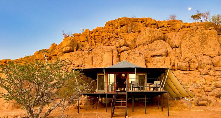 Scenic view of a tent-like cabin against dramatic rocks under a setting sun.