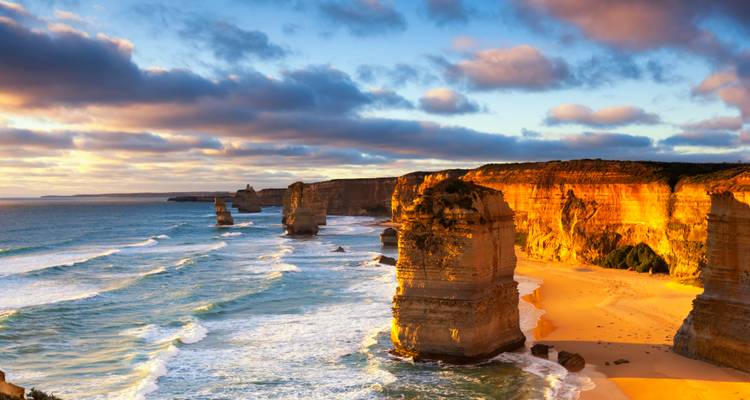 Pilas de piedra caliza iluminadas por el sol y acantilados erosionados de los Doce Apóstoles se alzan desde el turbulento Océano Austral bajo un cielo parcialmente nublado.