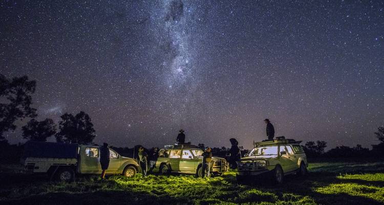 Grupo de viajeros con vehículos 4x4 acampan bajo un cielo nocturno deslumbrante lleno de estrellas en un entorno de pastizales del interior.