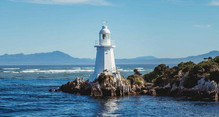 Faro blanco encaramado en un islote rocoso rodeado de un mar azul profundo con montañas a lo lejos.