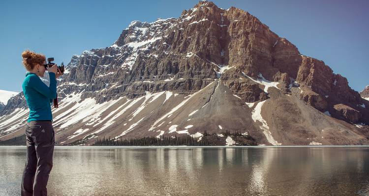 Viajera fotografiando una cresta de las Montañas Rocosas nevadas reflejada en un lago tranquilo.