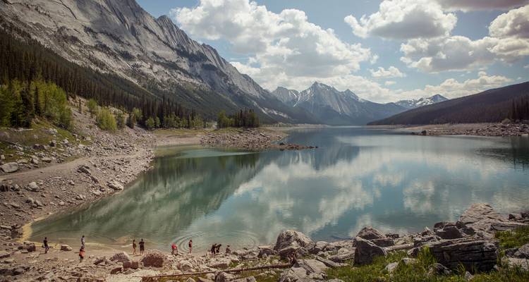 Pequeños grupos exploran la costa rocosa de un embalse alpino de aguas turquesas rodeado de montañas escarpadas.