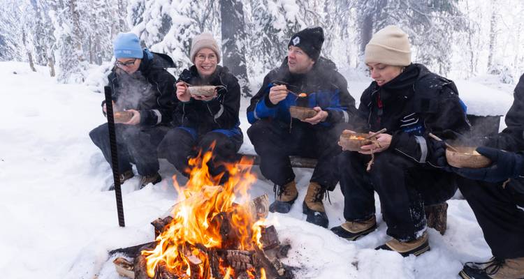Reisende wärmen sich an einem knisternden Lagerfeuer im verschneiten Wald auf und essen Suppe aus Holzschalen.