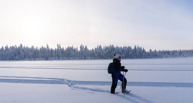 Einzelner Schneeschuhwanderer überquert breiten gefrorenen See unter heller Wintersonne mit Nadelwald am Horizont.