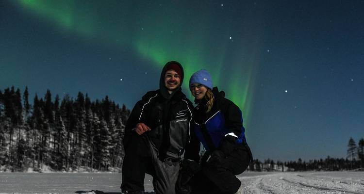 Pareja sonriente se arrodilla sobre el hielo de un lago nevado bajo una brillante aurora boreal verde en el cielo nocturno finlandés.