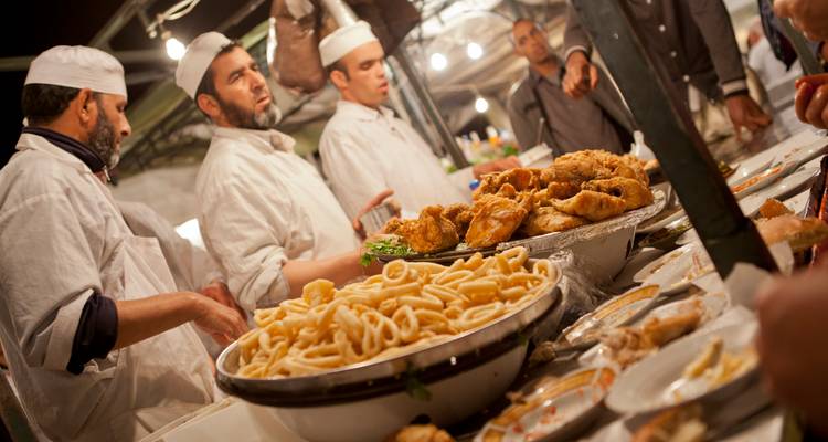 Des chefs de cuisine de rue en blouses blanches préparant une variété de plats marocains à un étal animé du marché nocturne.