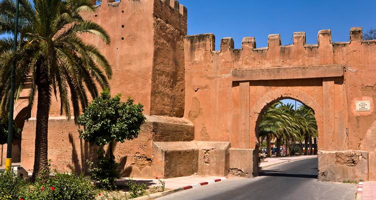 Porte fortifiée ensoleillée avec créneaux, palmiers et ciel bleu le long d'une voie de circulation.