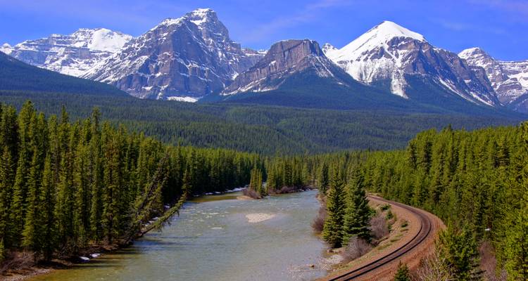 Curving railway track alongside a flowing river framed by dense evergreen forest and towering Rocky peaks.
