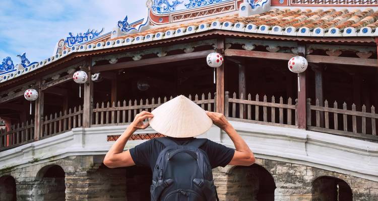 Een persoon die naar een traditionele Japanse brug kijkt terwijl hij een kegelvormige hoed draagt.