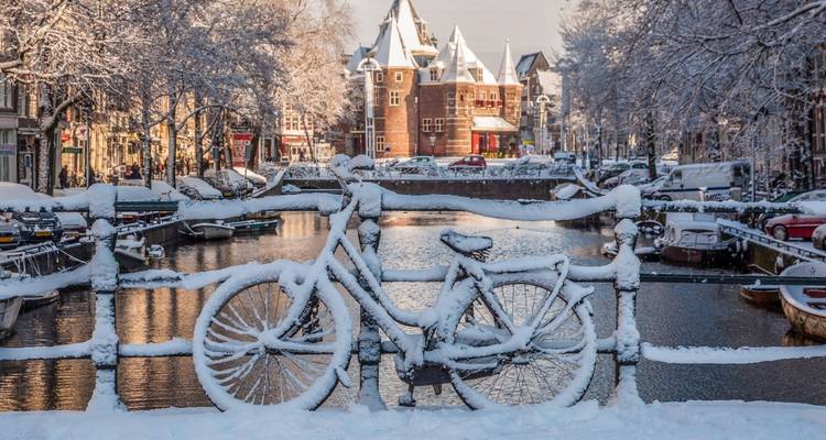 Vélo et rambarde recouverts de neige surplombant un canal hivernal d'Amsterdam et des bâtiments historiques.