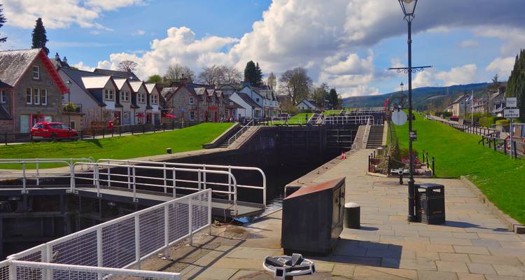 Kleurrijke dorpshuizen langs een zonnige kanaalsluissluis met groene heuvels op de achtergrond