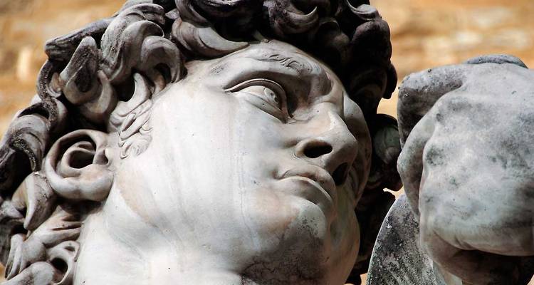 Vista de cerca de la estatua de mármol David de Miguel Ángel mostrando la cara detallada y los rizos contra un fondo de piedra cálida.