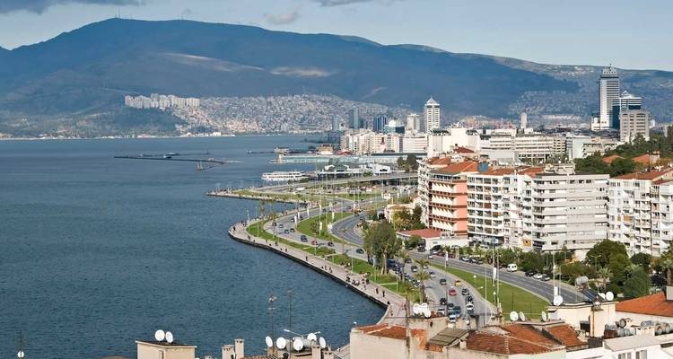Panoramische Küstenstadtansicht von Izmir mit einer geschwungenen Uferpromenade, Uferparks, modernen Gebäuden und fernen Bergen über der blauen Bucht.