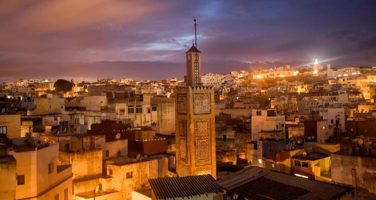Vista del atardecer sobre una antigua ciudad del norte de África con un minarete decorado con azulejos que se alza sobre edificios de tonos terrosos densamente agrupados bajo un cielo vespertino colorido.