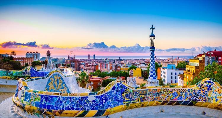 Otro ángulo de la terraza de mosaicos icónica del Park Güell con tonos de amanecer sobre Barcelona.