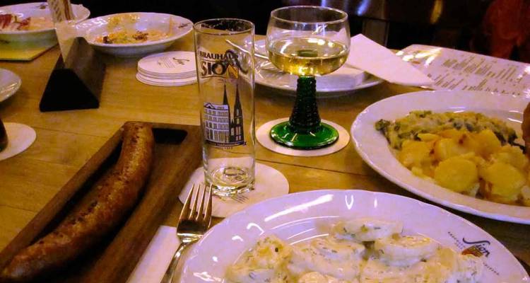 Comida alemana rústica en una mesa de madera con salchicha, platos de papa y cristalería de cerveza con la marca Köln.