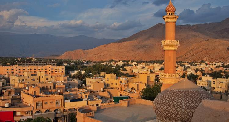 Una luz dorada baña un minarete y una mezquita de cúpula que domina el pueblo desértico de Nizwa con montañas al fondo.
