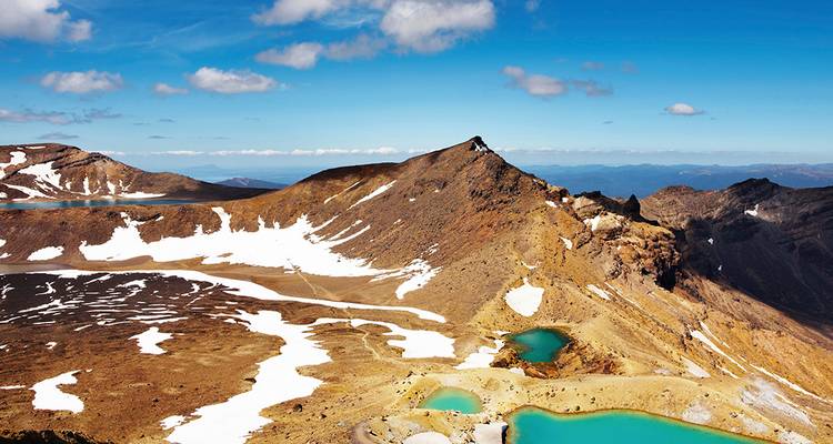 Paysage alpin volcanique avec des lacs de cratère turquoise, des plaques de neige et une crête abrupte sous un ciel dégagé.