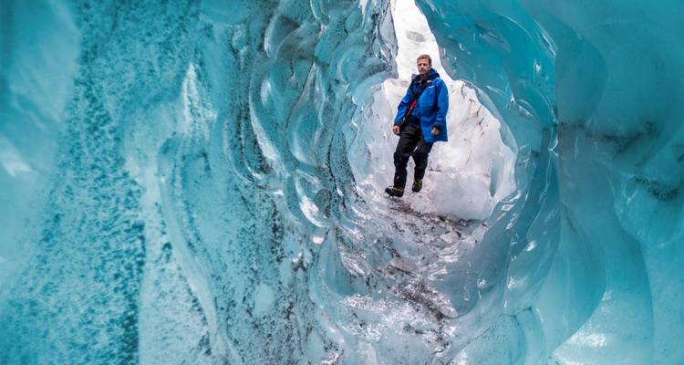 Un voyageur explore un tunnel de glace bleu scintillant à l'intérieur d'un glacier, entouré de parois sculptées.