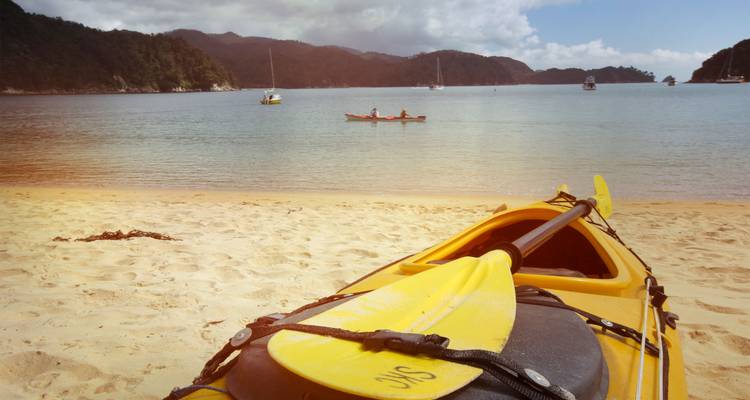 Un kayak de mer échoué avec une pagaie jaune repose sur le sable doré face à une baie tranquille parsemée de bateaux.