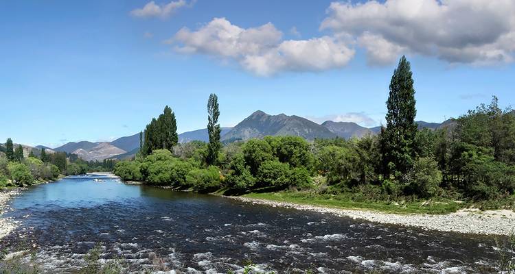 Une rivière cristalline s'écoule à travers une large vallée verdoyante avec des montagnes qui s'élèvent au loin sous un ciel ensoleillé.