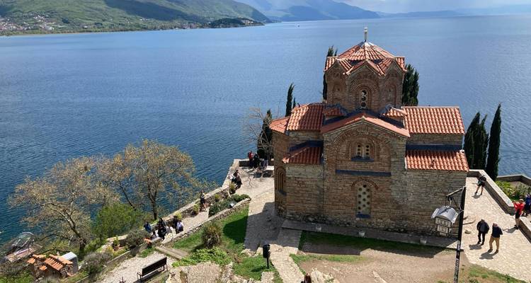 Iconische kerk van Sint-Jan te Kaneo die hoog boven de diepblauwe wateren van het Meer van Ohrid uittorent, met bezoekers die in de buurt slenteren.