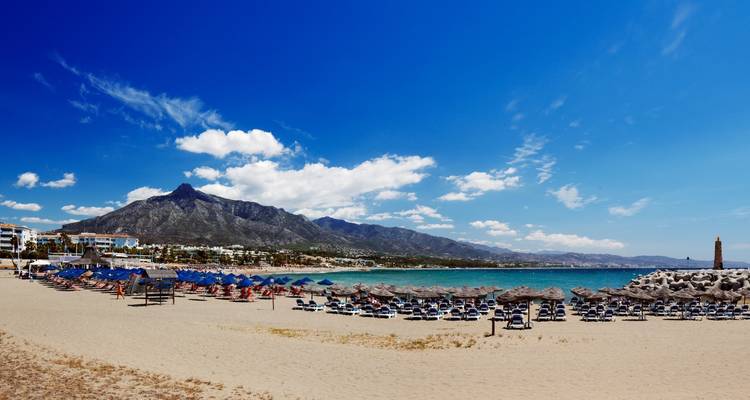 Breed zandstrand met rijen blauwe ligbedden en parasols met uitzicht op turquoise water en bergen in de verte.