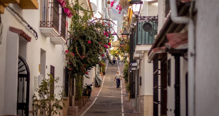 Bloemversierde smalle steeg in een witgekalkt Andalusisch dorpje met een enkele voetganger die omhoog loopt.