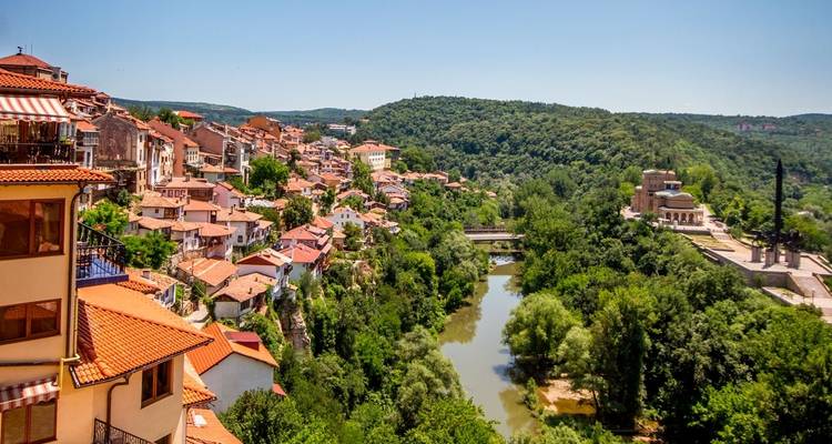Panorama escénico de los tejados de terracota de Veliko Tarnovo bordeando un desfiladero fluvial rodeado de colinas boscosas.