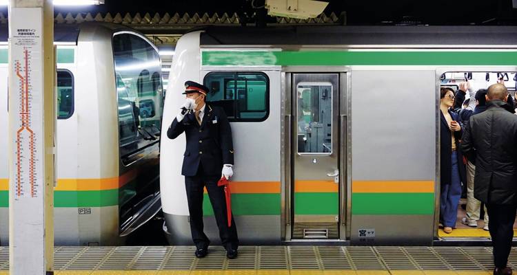 Un contrôleur de train japonais se tient sur le quai à côté d'un train de banlieue argenté et vert pendant que les passagers montent à bord.