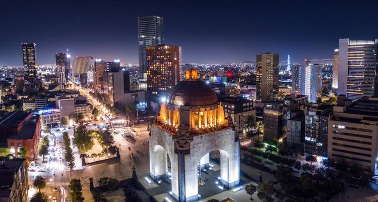 Vista aérea dramática nocturna de la Ciudad de México mostrando el Monumento a la Revolución iluminado entre rascacielos modernos.
