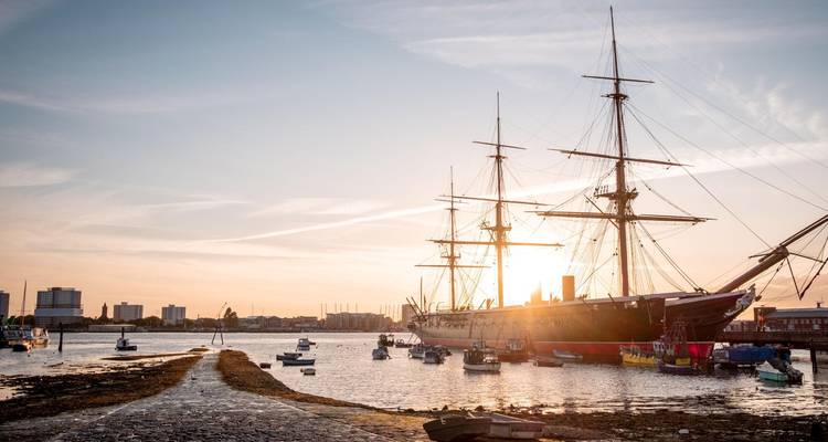 Velero histórico anclado en un puerto con el perfil de la ciudad recortado contra un cálido cielo al atardecer.