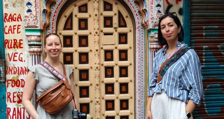 Dos viajeros están parados junto a una puerta de madera ornamentada y pintada de colores brillantes en un callejón indio.