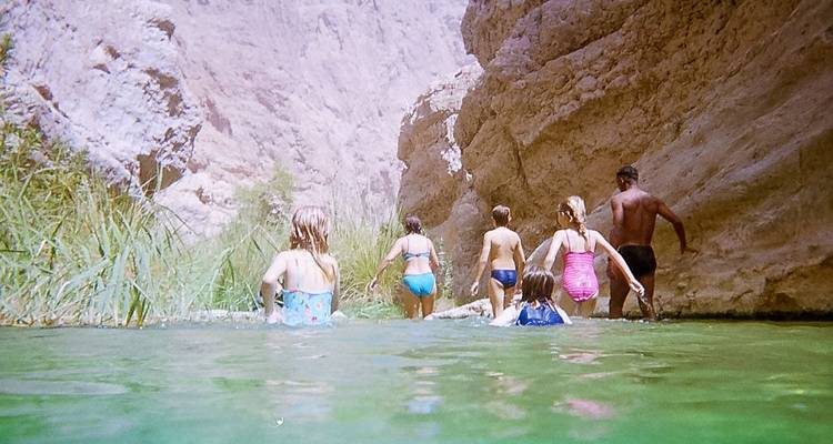 Adultos y niños caminan por agua esmeralda en un estrecho cañón rocoso de wadi.