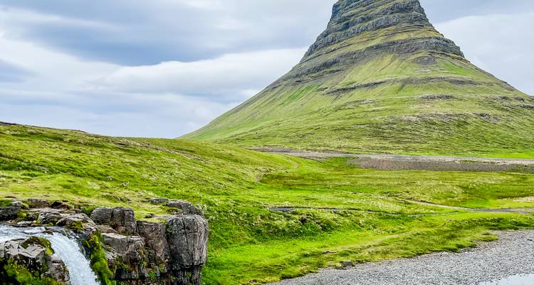 Montagne verte iconique en forme de cône s'élevant derrière une petite cascade et un paysage herbeux luxuriant sous un ciel nuageux.