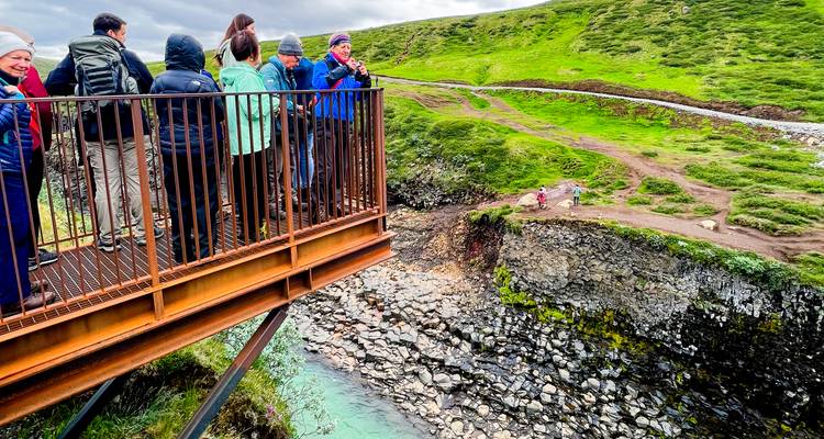 Un groupe de touristes vêtus de vêtements de plein air se tient sur une plateforme d'observation en métal surplombant une gorge rocheuse avec de l'eau turquoise et des collines verdoyantes.