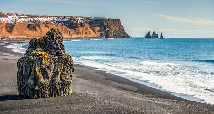 Plage de sable noir spectaculaire avec un rocher isolé couronné de mousse et des falaises de basalte lointaines s'élevant de l'Atlantique bleu éclatant.