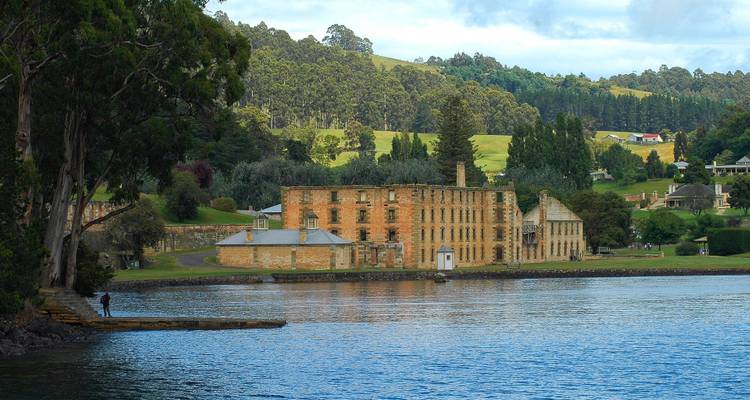 Les bâtiments historiques en brique du pénitencier de Port Arthur encadrés par de grands arbres et les eaux calmes d'une baie.