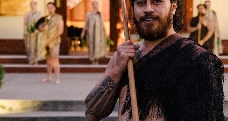 Intérprete maorí en vestimenta tradicional con tatuaje facial y bastón de madera tallado durante una ceremonia cultural.