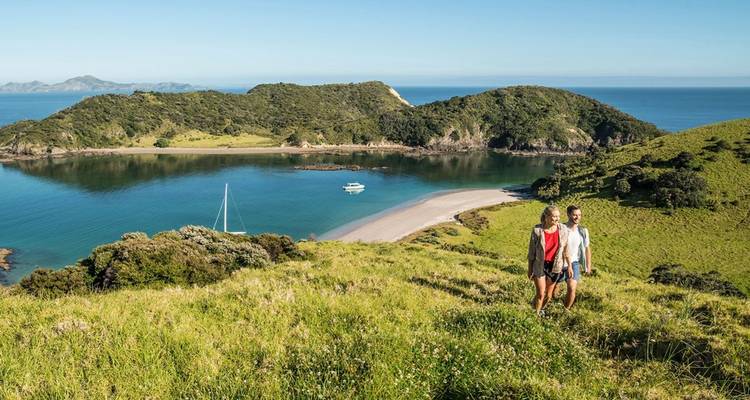 Pareja caminando por una cresta cubierta de hierba con vista a una bahía turquesa tranquila y promontorios verdes.