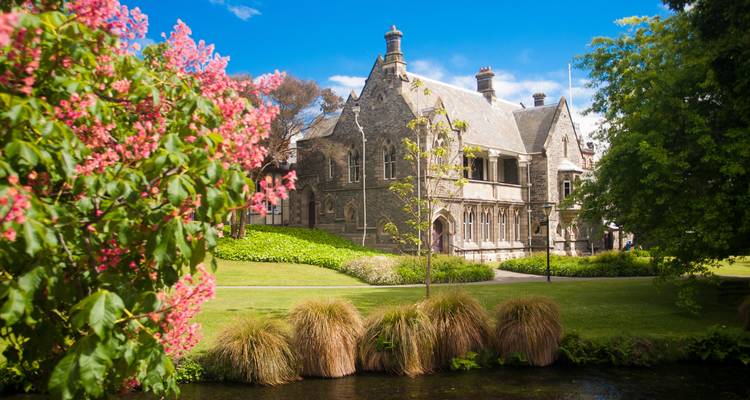Edificio histórico de piedra enmarcado por árboles en flor, césped bien cuidado y un arroyo tranquilo en Christchurch.