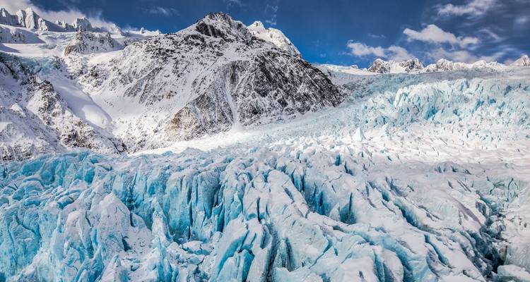 Las grietas azules irregulares dividen el vasto hielo del Glaciar Franz Josef mientras los picos cubiertos de nieve se elevan detrás.